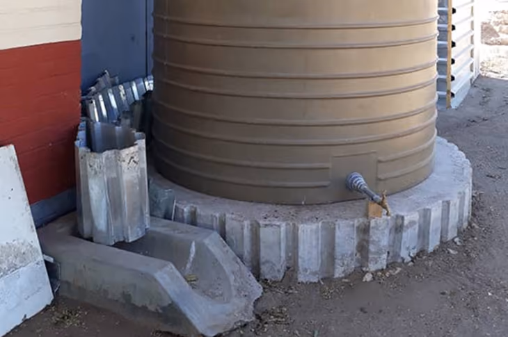 Brauner runder Wassertank mit Metallhahn auf Betonbasis neben einem kleinen Entwässerungsgraben.