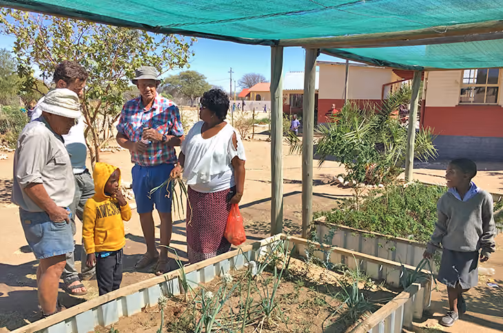 Gruppe von Erwachsenen und Kindern im schattigen Gemüsegarten vor einer Grundschule.