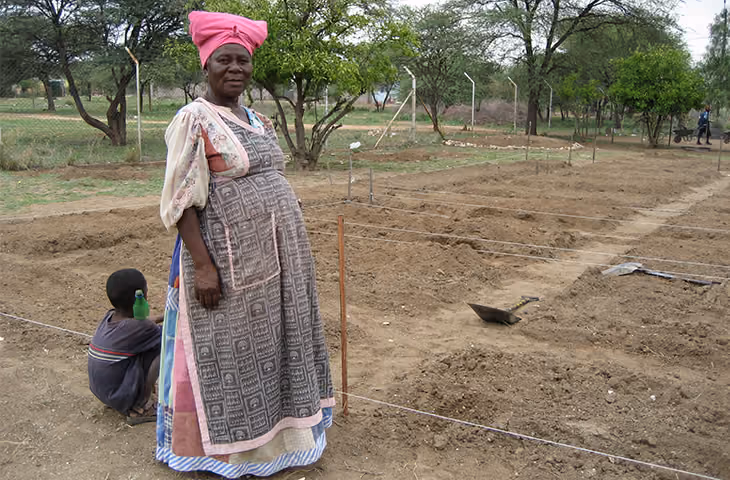 Frau in traditioneller Kleidung steht in einem präparierten Garten mit einem Kind, das in der Nähe sitzt.