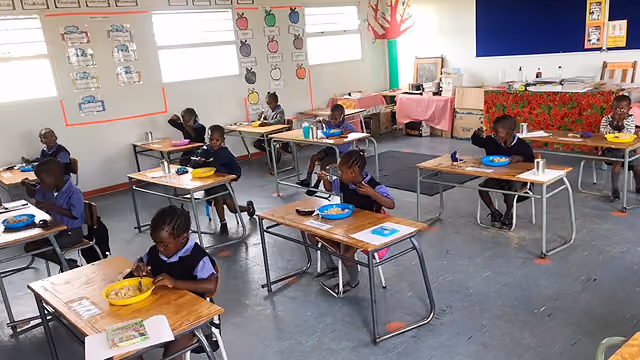 Grundschulkinder sitzen einzeln an Tischen in einem Klassenraum und essen aus bunten Schalen.