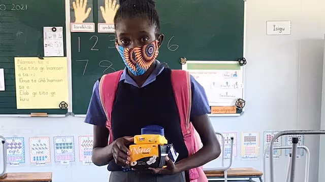 Schülerin mit buntem Maskenstoff hält eine gelbe Brotdose in einem Klassenzimmer.