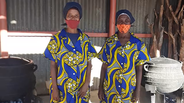 Zwei Frauen in bunten Kleidern und Gesichtsmasken in einer Küche mit großen Töpfen.
