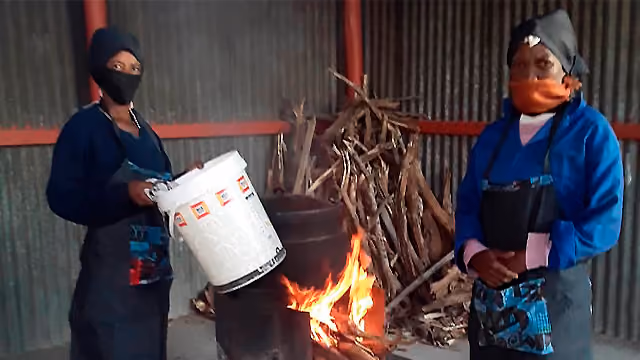 Two women wearing masks stand near a barrel fire with firewood inside a metal structure.