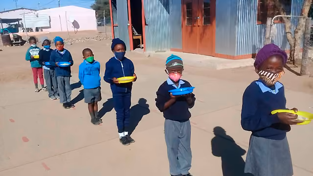 Schulkinder mit Masken stehen in einer Reihe im Freien und halten bunte Teller in der Hand.