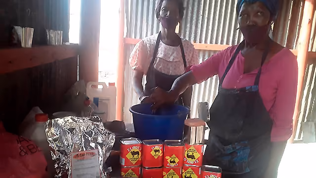Two women wearing masks and aprons preparing food behind stacked red tin cans in a rustic setting.