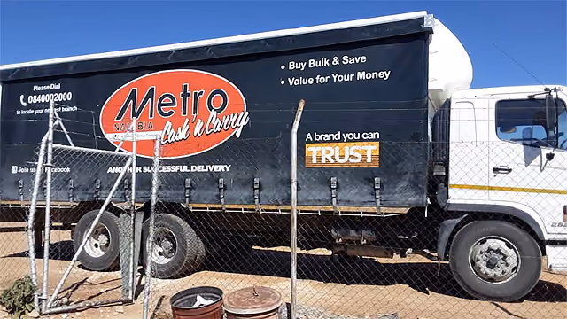 Lkw mit Werbung für Metro Namibia im Freien hinter einem Maschendrahtzaun bei klarem Himmel.