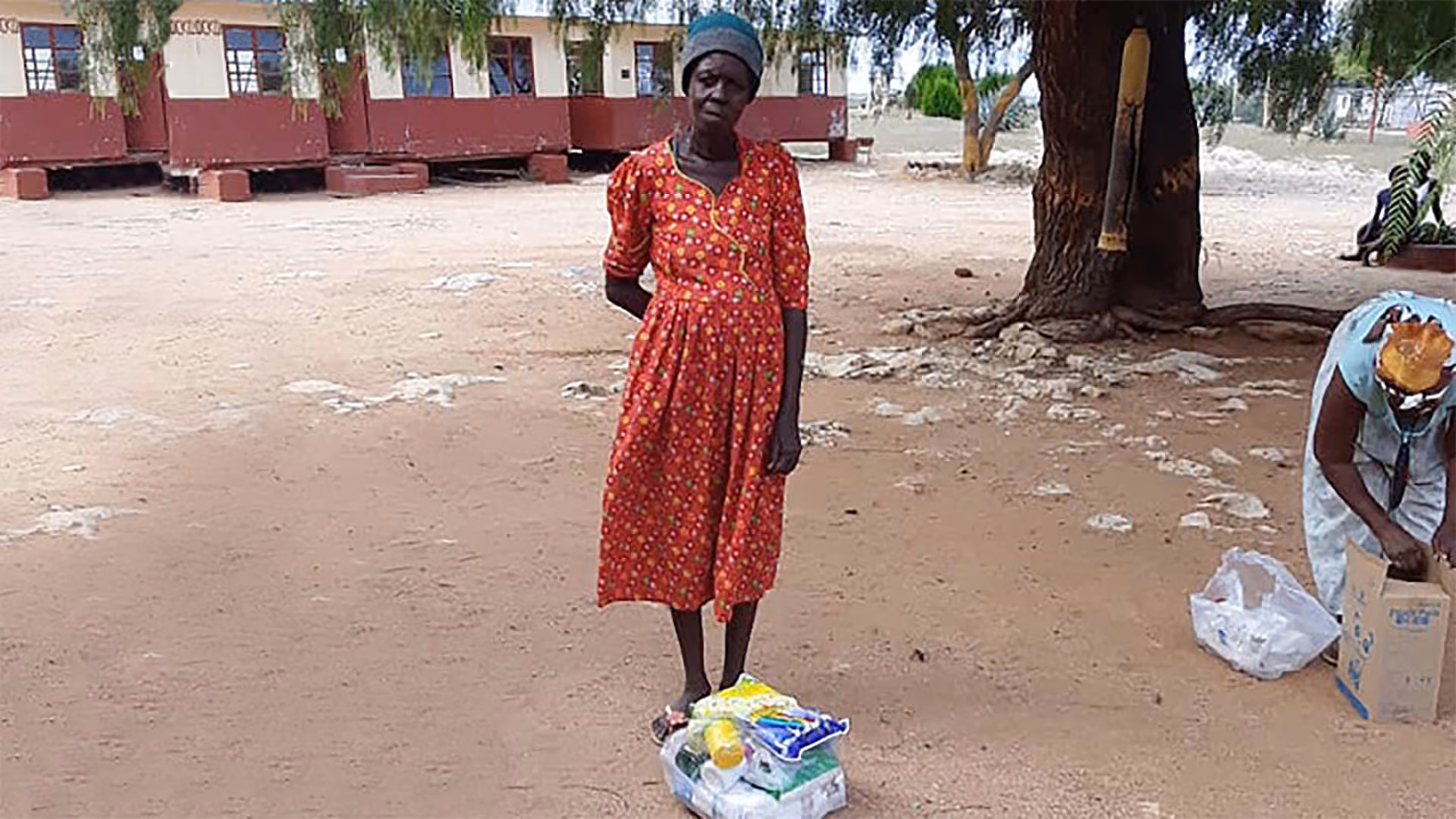 Frau in rotem Kleid mit Lebensmitteln in Plastiktüte auf sandigem Boden vor einem Baum und Gebäude.