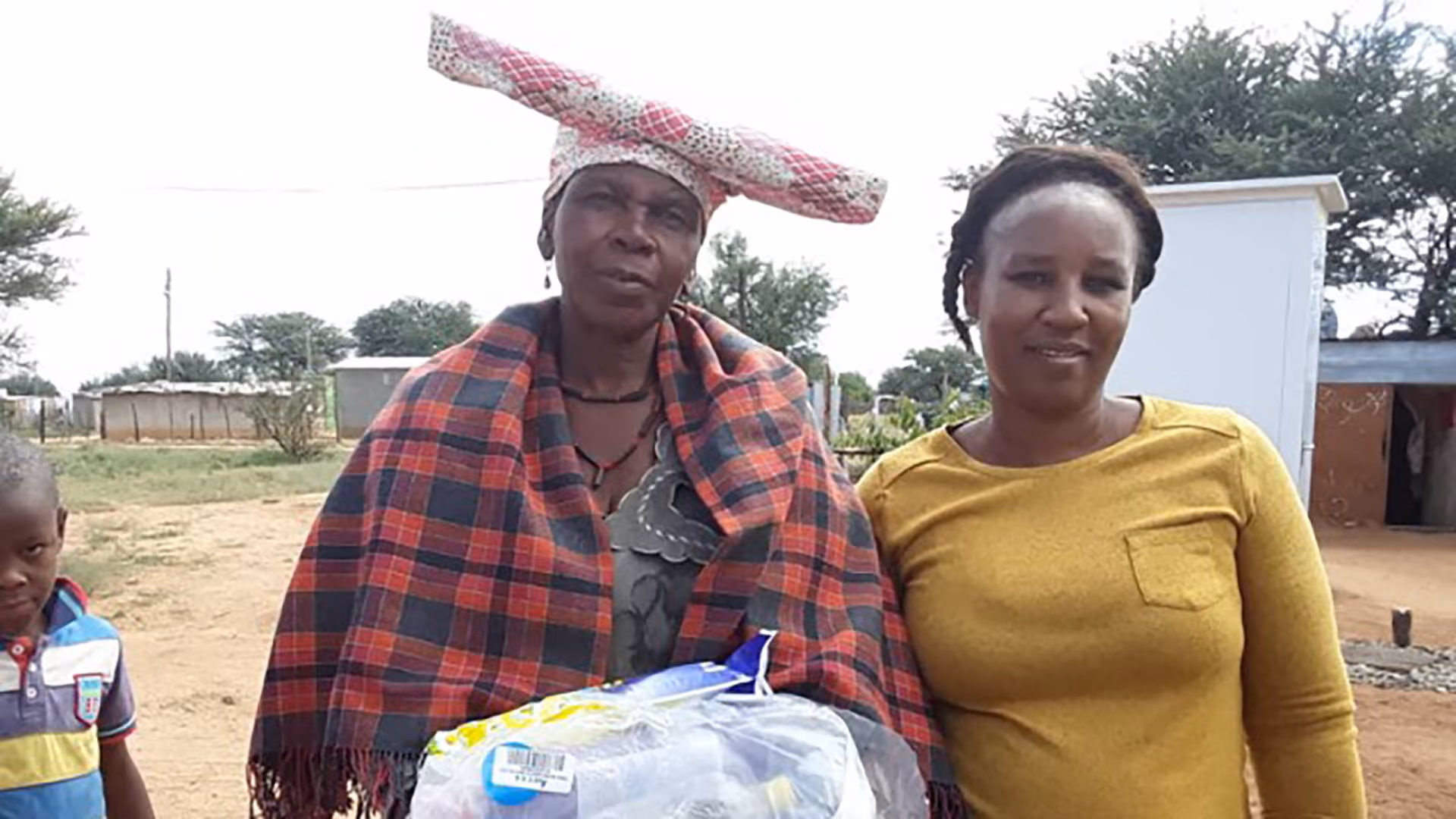 Zwei afrikanische Frauen, eine mit buntem Kleid und Kopfbedeckung, stehen im Freien, ein Kind dabei.