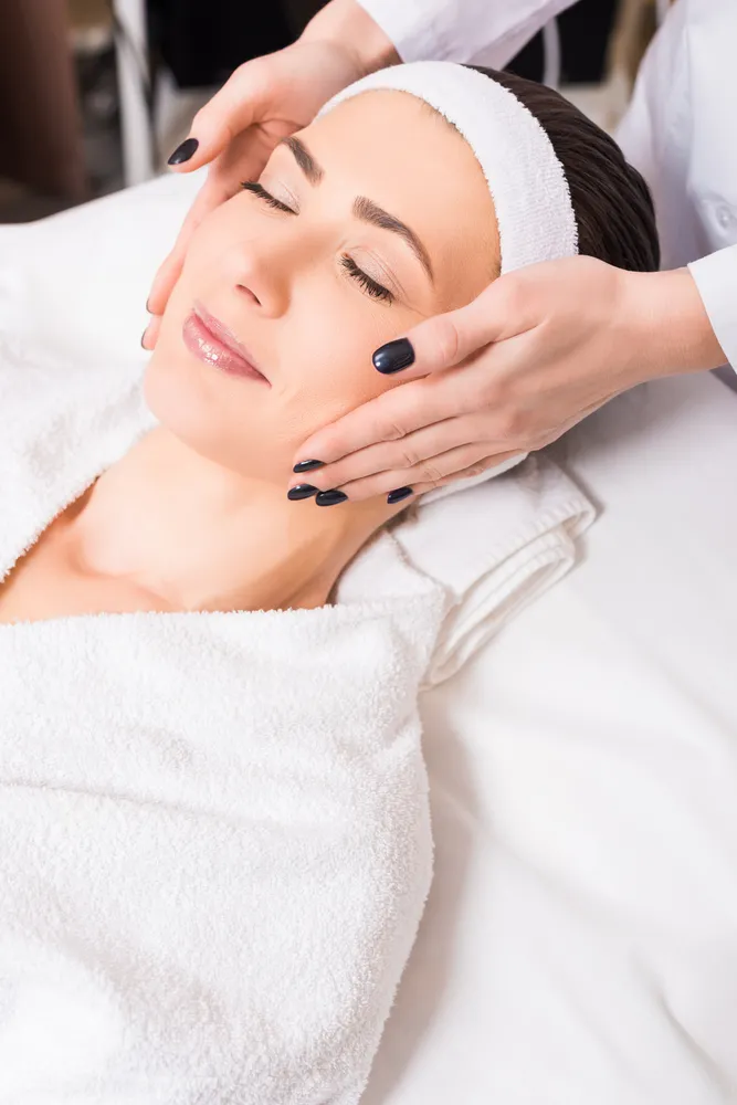 Woman with eyes closed receiving a facial massage while lying on a spa bed, wearing a white headband and robe.