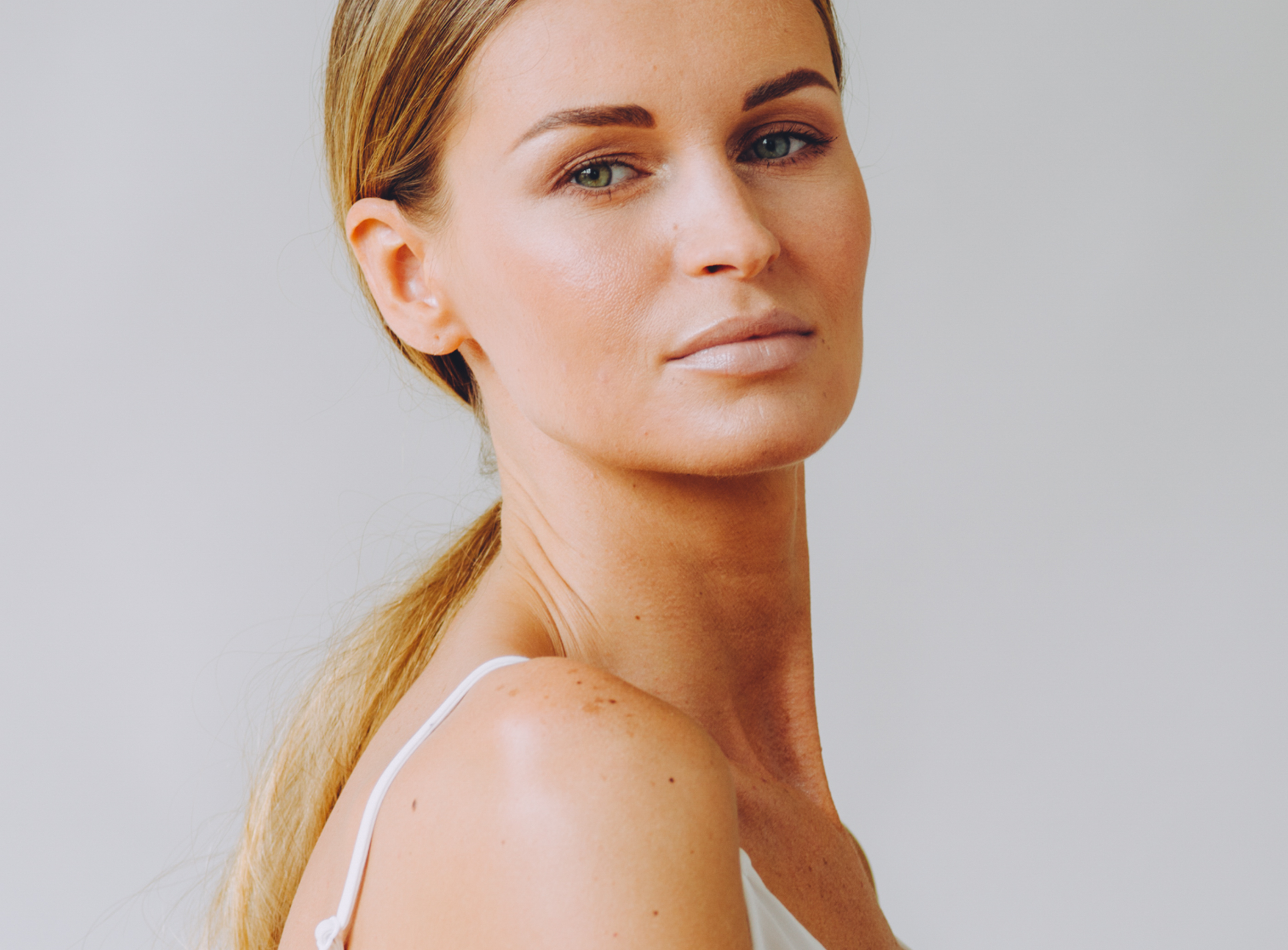 Close-up portrait of a woman with light brown hair tied back, wearing a white sleeveless top against a plain light background.