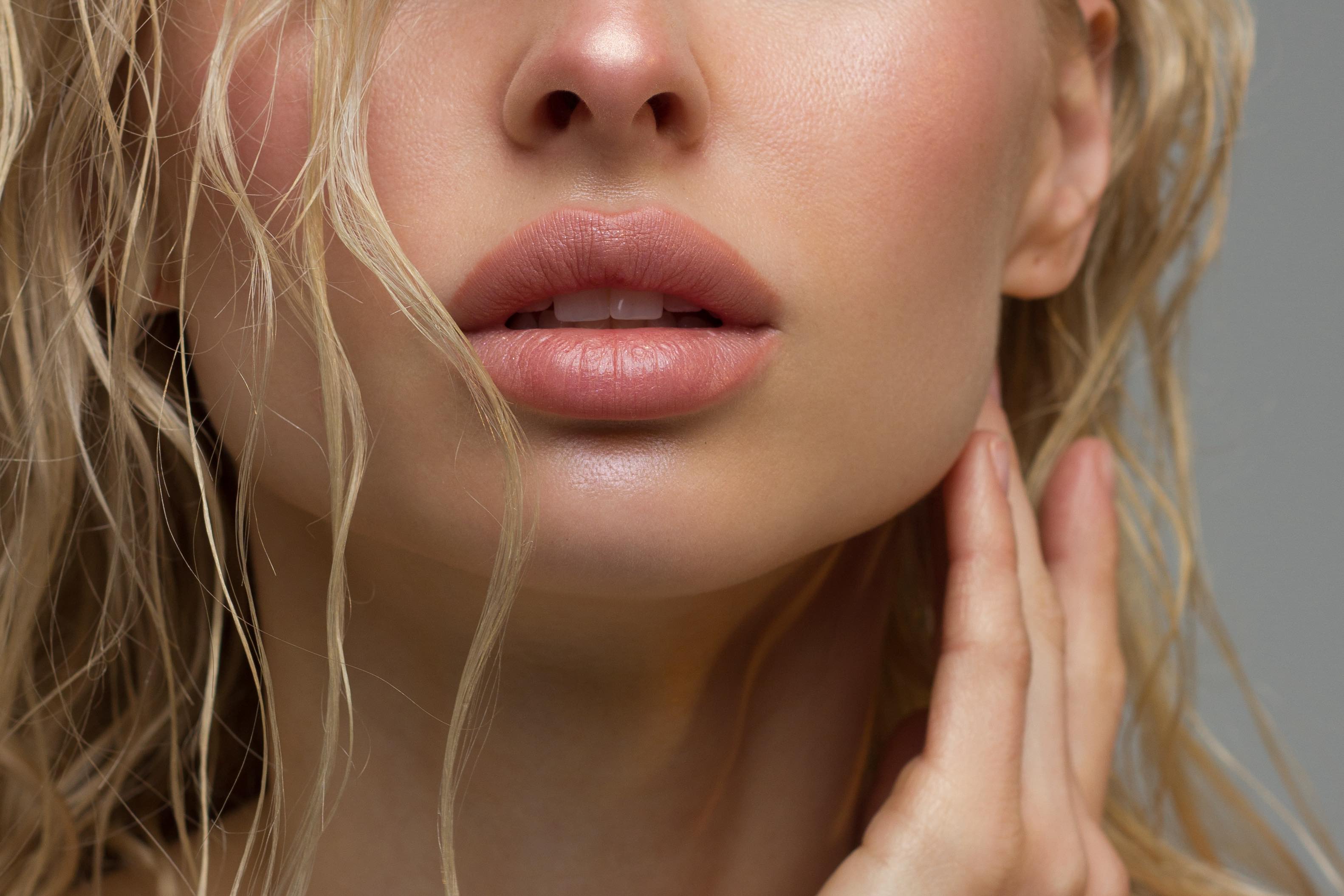 Close-up of a woman's lips, chin, and nose with wavy blonde hair strands and hand gently touching her neck.