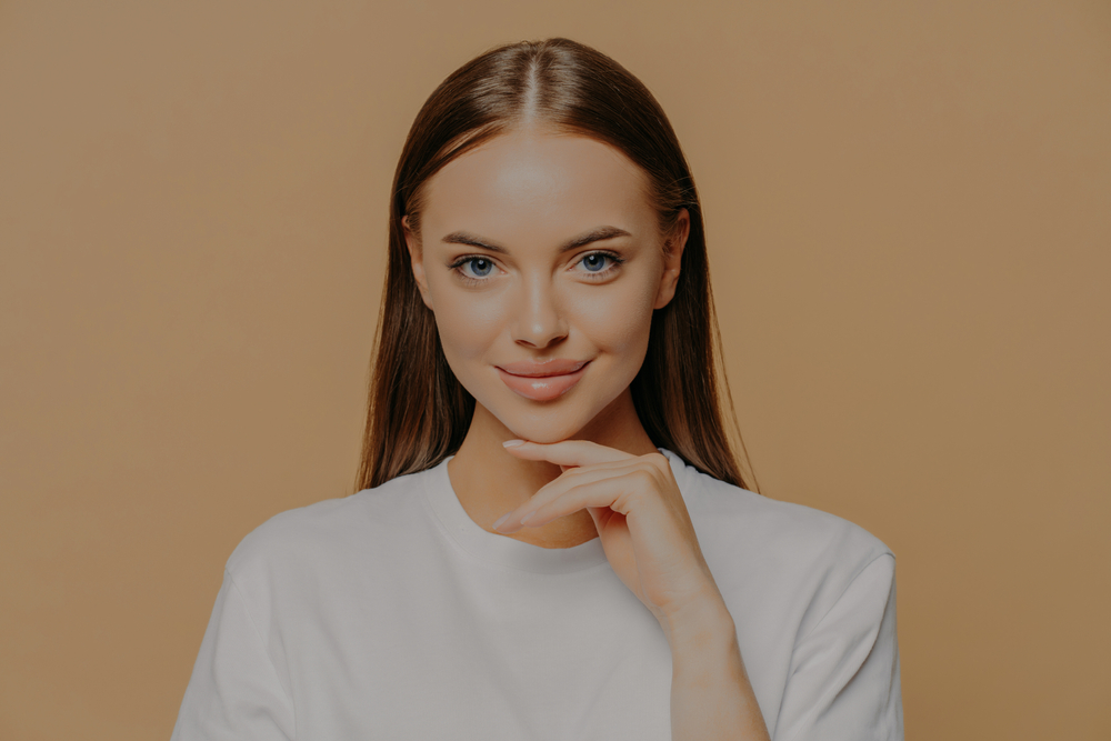 Smiling young woman with long brown hair and blue eyes wearing a white shirt against a beige background.