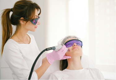A professional wearing pink gloves performs a laser skin treatment on a reclining woman wearing protective glasses.