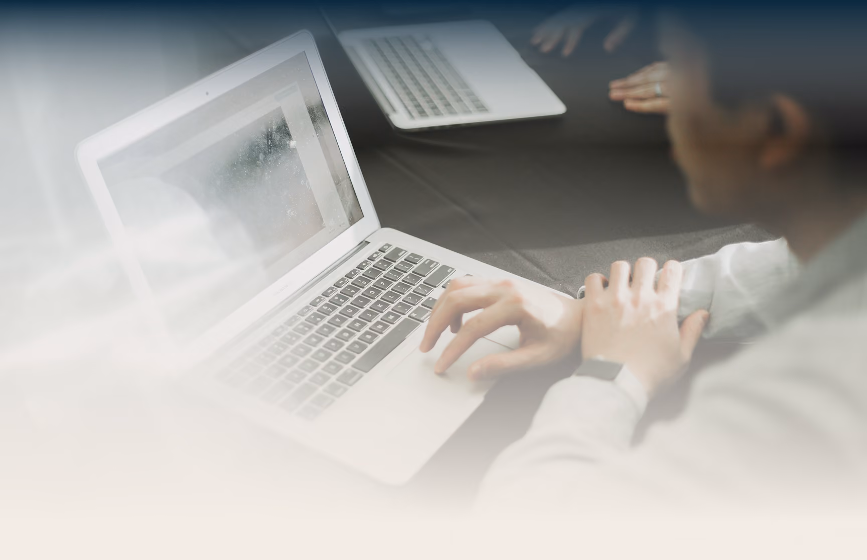 Hände tippen auf einem Laptop, daneben ein zweites Notebook am Tisch