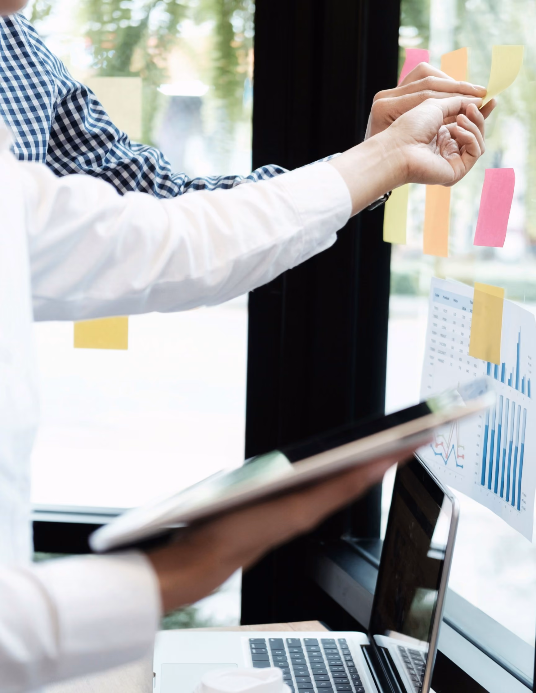 Person heftet Haftnotizen an eine Glasscheibe mit Diagrammen, daneben ein Tablet