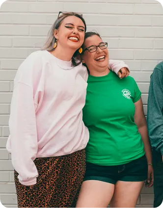 Two women smiling and hugging in front of a white brick wall, one in a pink sweatshirt, the other in a green shirt.