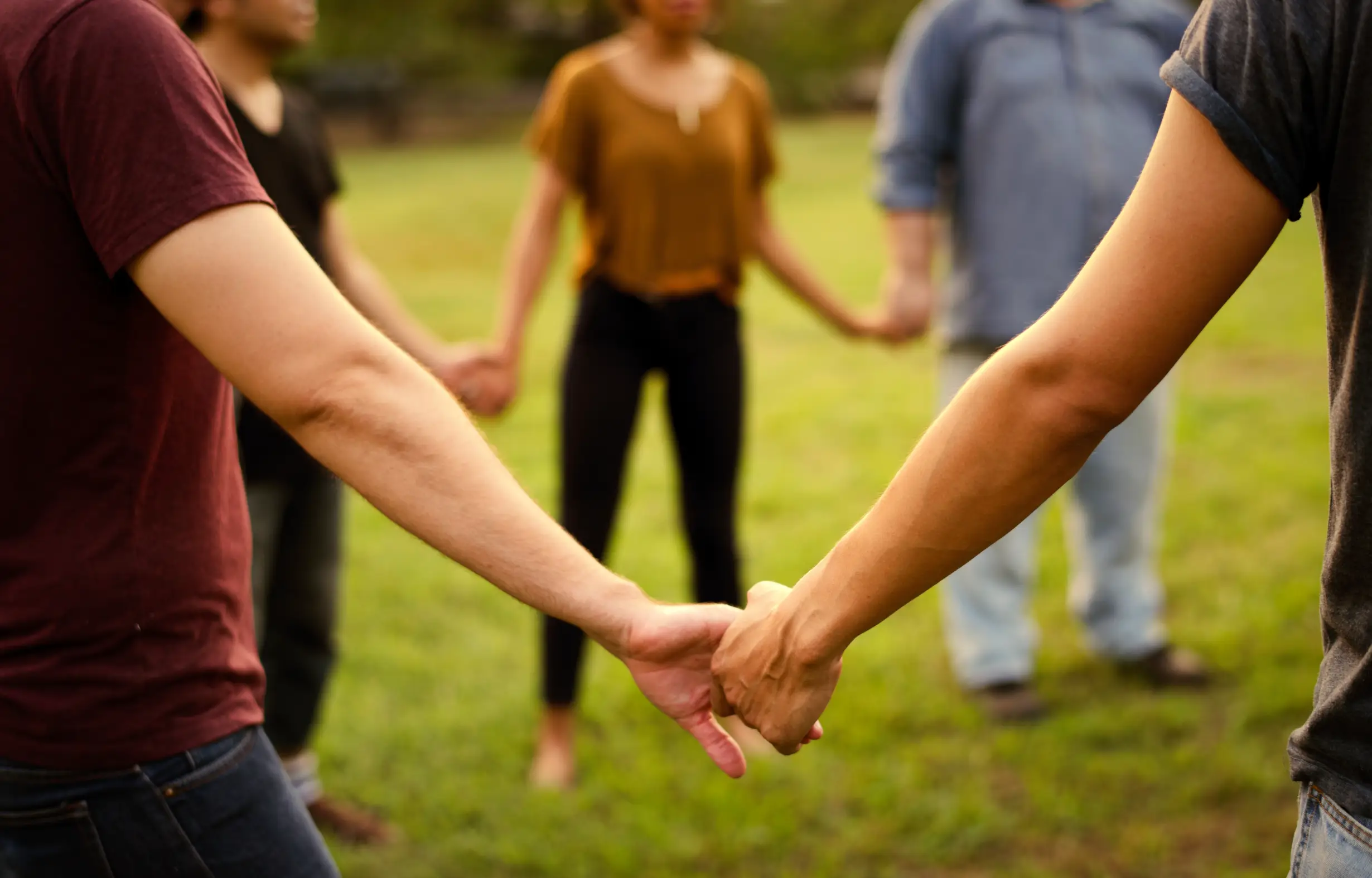 Group of people standing in a circle holding hands outdoors on grass.