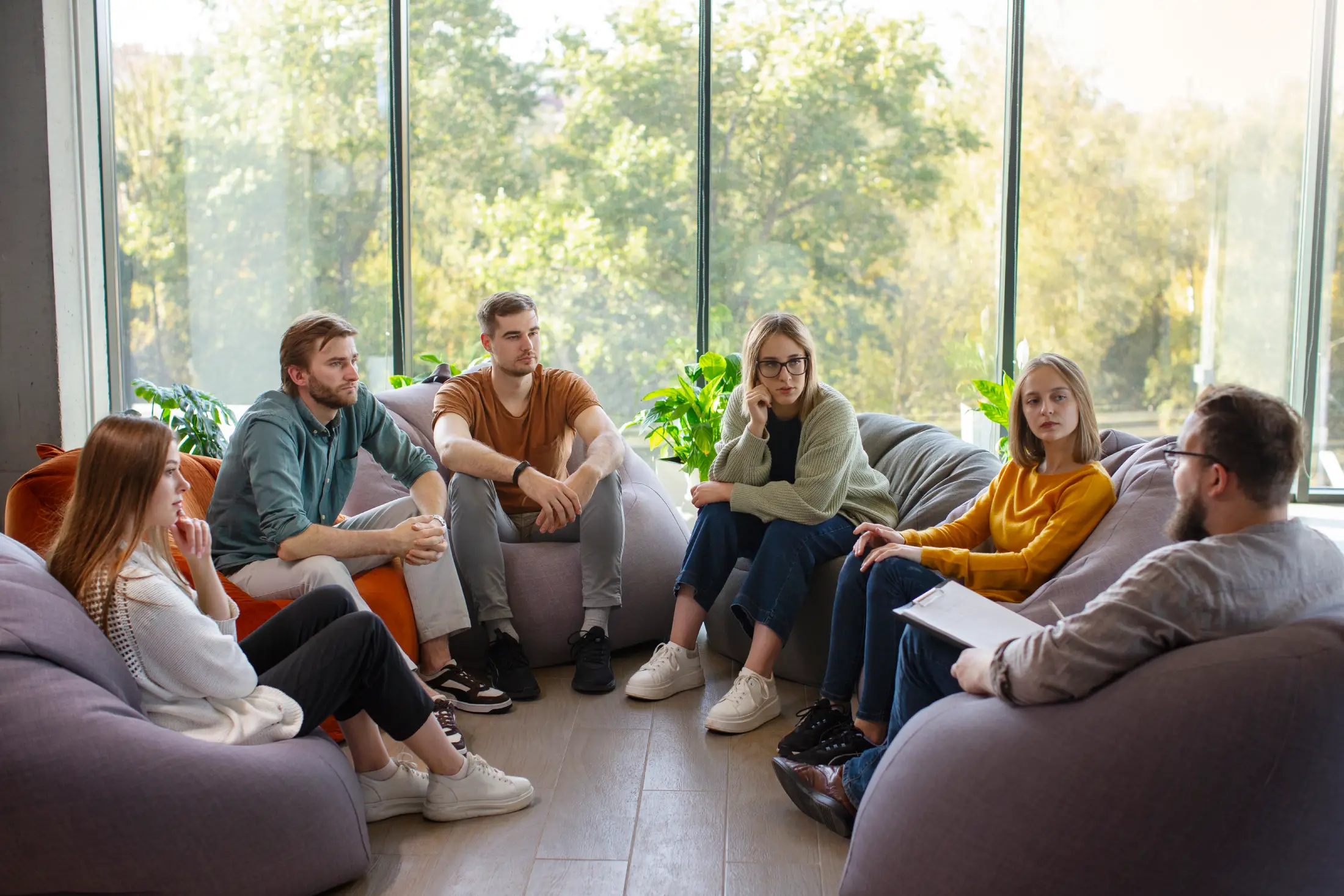 Group therapy session with five people seated in a circle on cushioned chairs in a bright room.