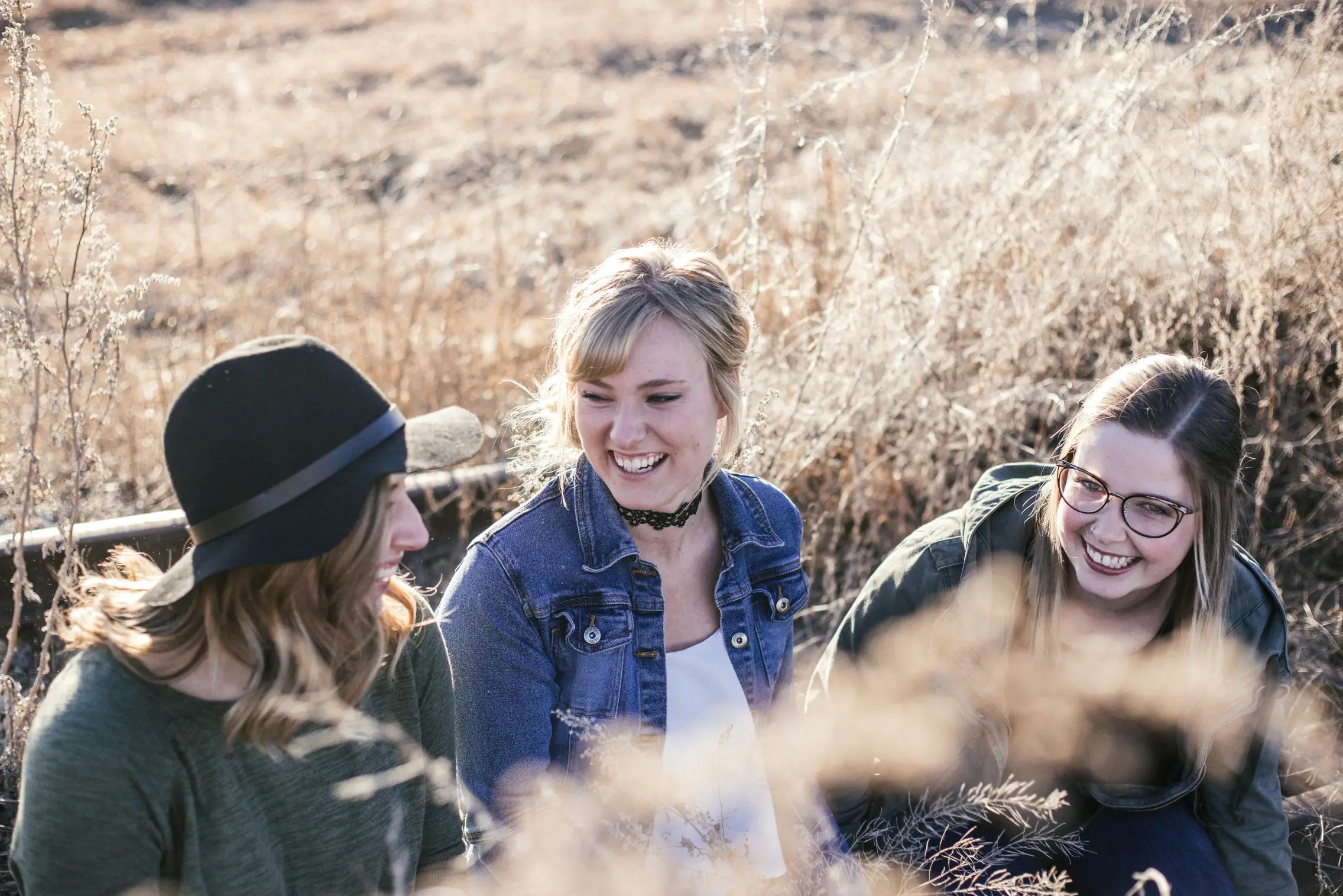 Three young women sitting outdoors in dry grass smiling and talking to each other.