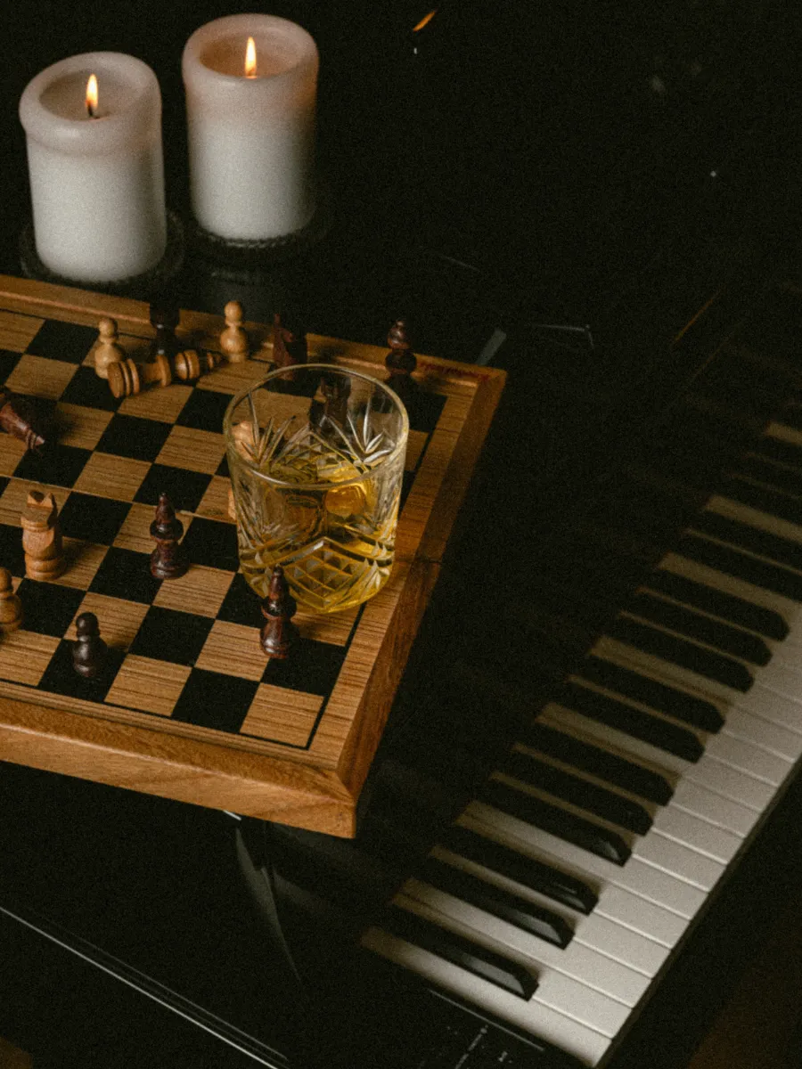 Verre de whisky posé au milieu d'un échiquier en partie, deux bougies allumées éclairant le clavier de piano en arrière-plan