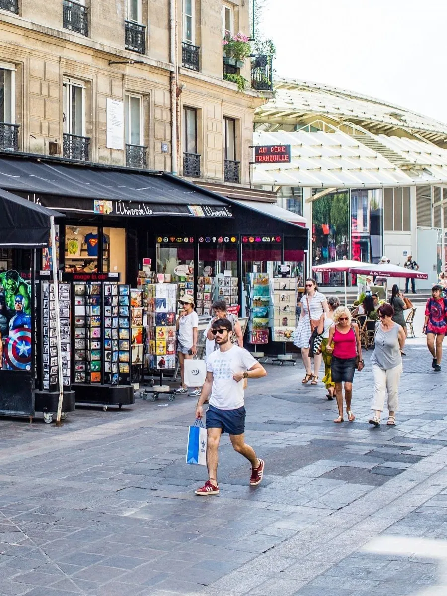 Kiosque de cartes postales et souvenirs sur une place pavée fréquentée, immeubles haussmanniens et verrière du marché en toile de fond