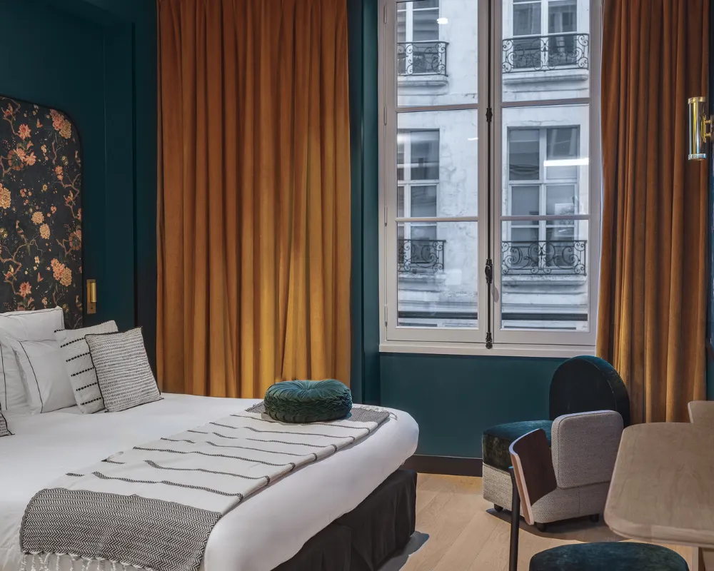 Chambre bleu paon avec lit à coussin vert rond, rideaux moutarde et fenêtre parisienne, fauteuils gris et vert en coin