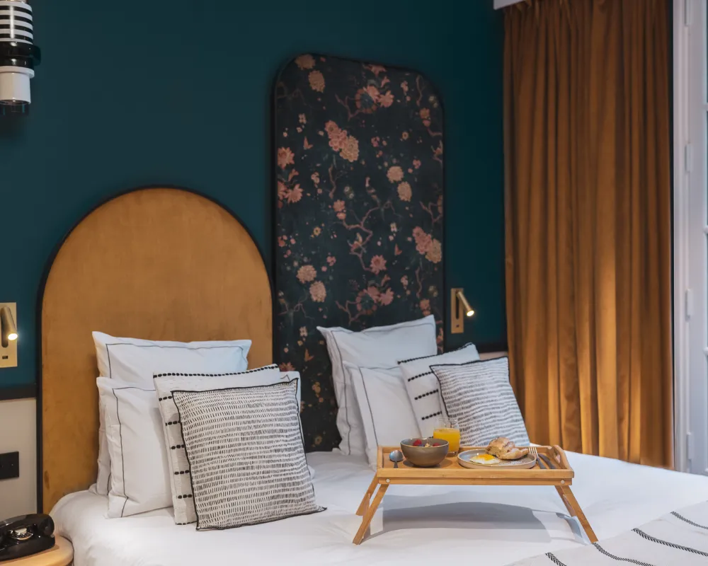 Deux lits jumeaux à têtes de lit dorée arquée et panneau floral, plateau petit-déjeuner posé au centre devant un rideau moutarde