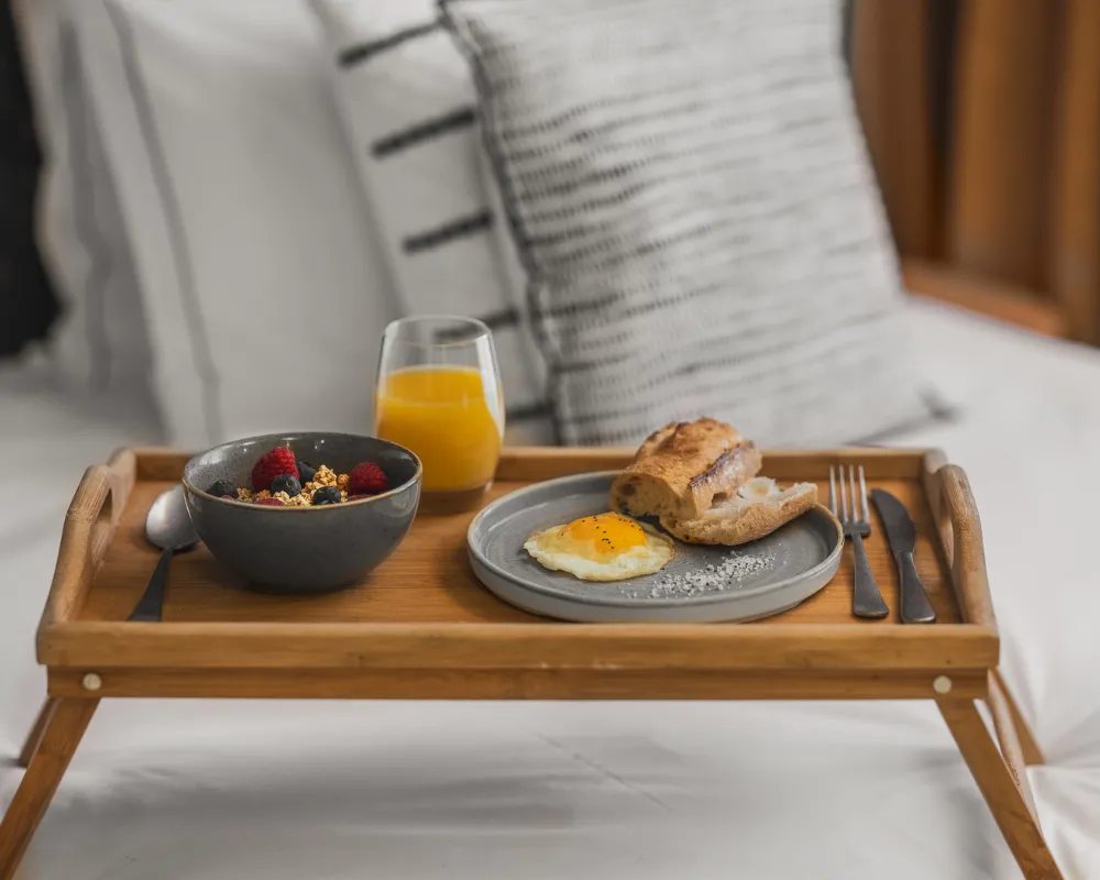 Plateau petit-déjeuner en bois avec bol de fruits rouges, jus d'orange et œuf au plat, posé sur un lit à coussins rayés