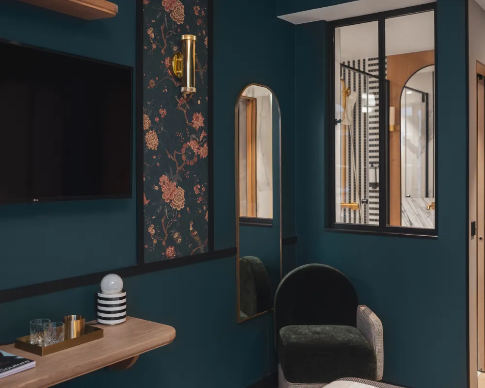 Coin bureau bleu paon avec télévision murale, miroir arqué doré et fauteuil noir devant une verrière donnant sur une salle de bain