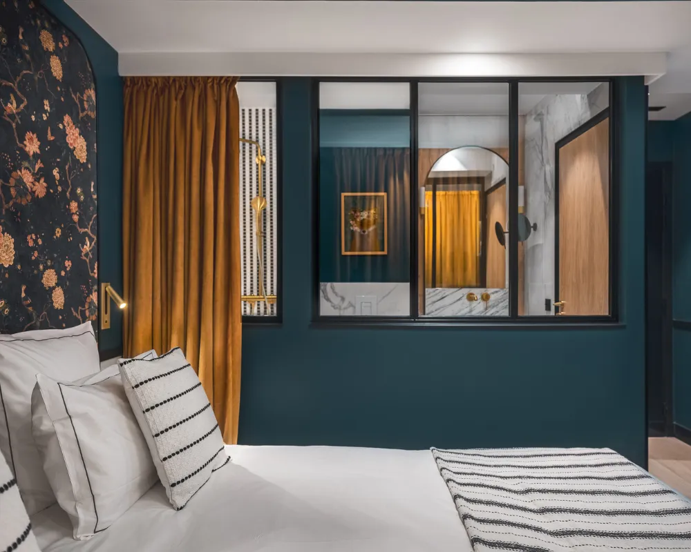 Lit à panneau floral sombre et rideau moutarde, verrière vitrée ouvrant sur une salle de bain éclairée en fond