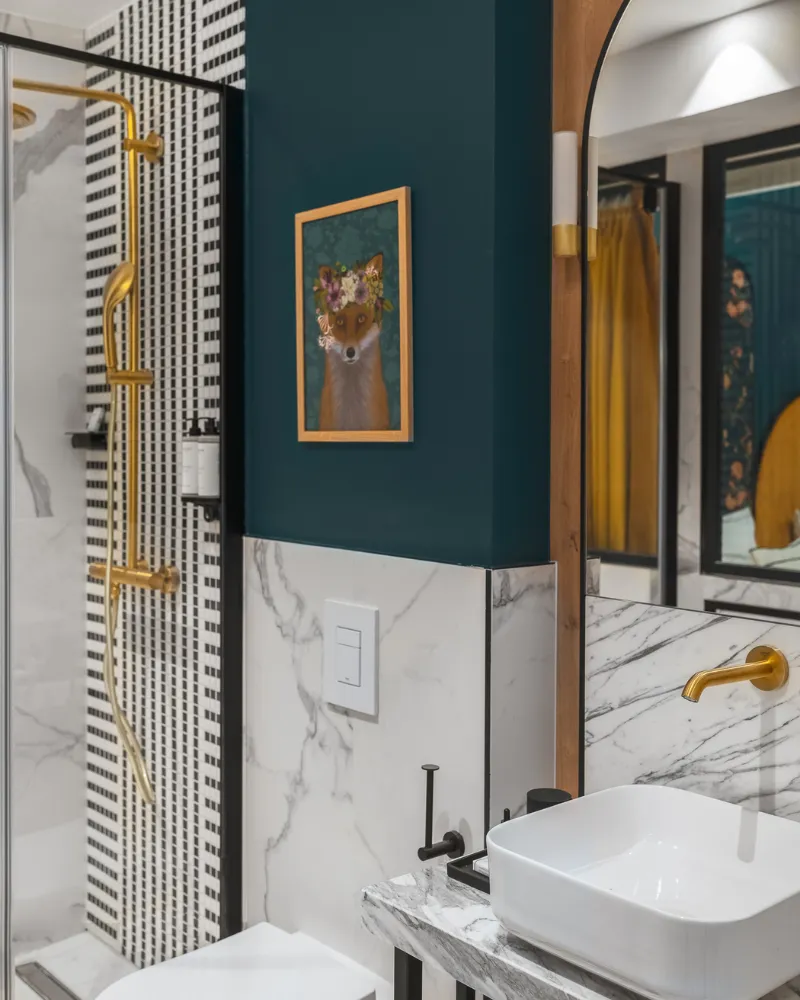 Salle de bain en marbre veiné avec douche à robinetterie dorée, tableau encadré doré et vasque à double poignée