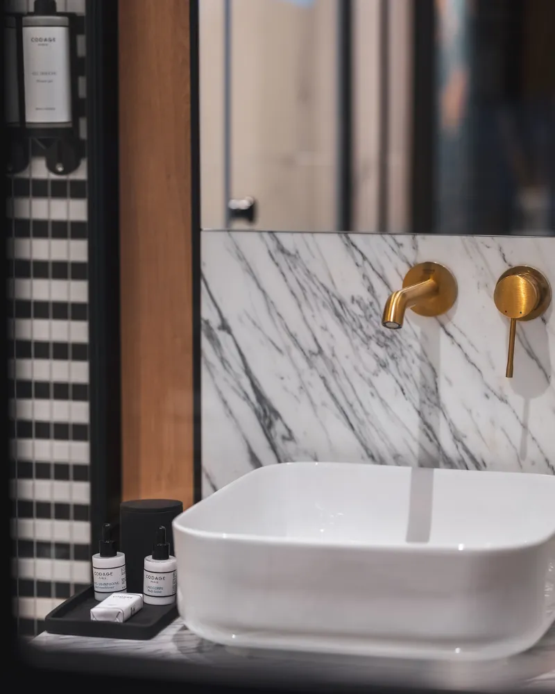 Vasque blanche à robinetterie dorée sur plan en marbre veiné, miroir et douche à mosaïque noire et blanche en fond