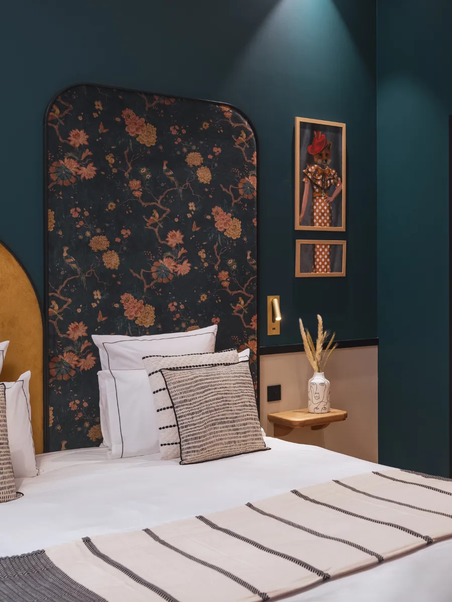 Tête de lit florale bordée de bleu paon, tableaux illustrés de mode encadrés en doré et vase en forme de visage sur table de chevet