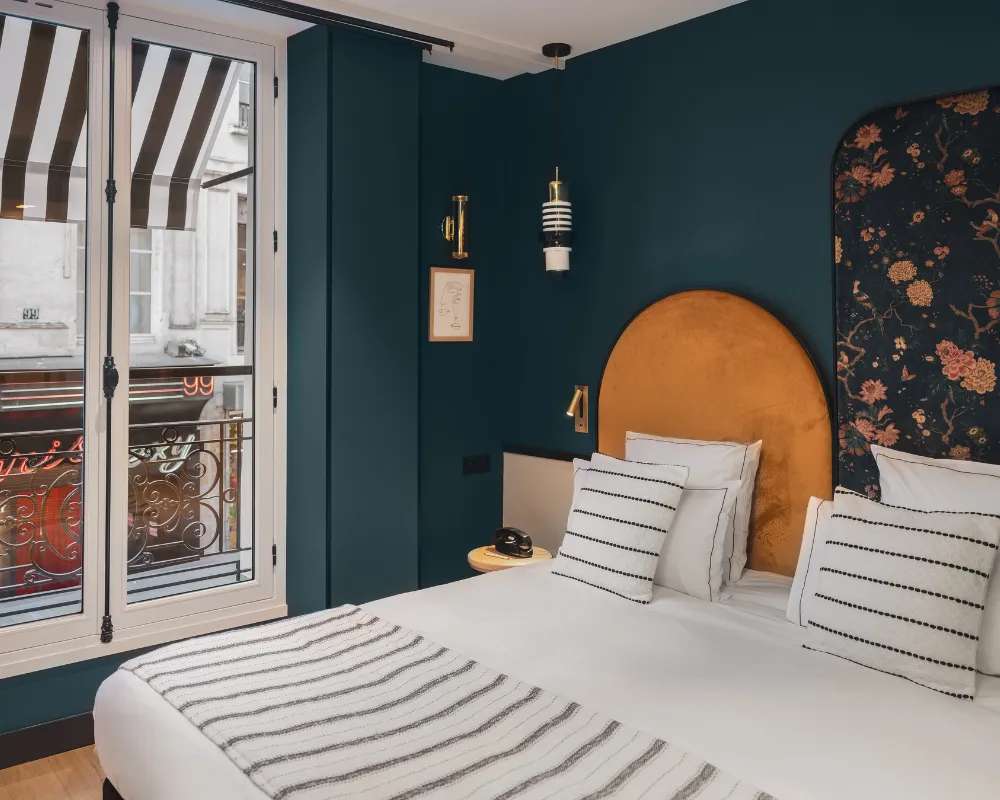 Chambre bleu paon avec tête de lit dorée arquée et panneau floral, fenêtre ouverte sur un balcon sous auvent rayé