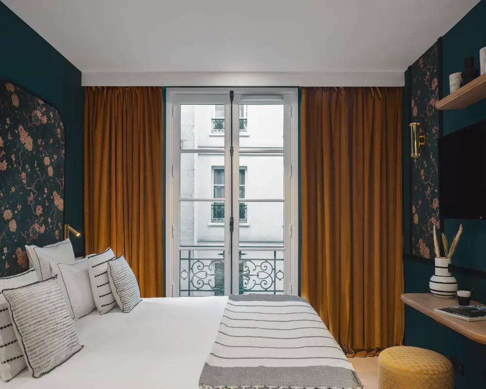Chambre bleu paon au papier peint floral orangé, rideaux moutarde et porte-fenêtre ouverte sur un balcon parisien, pouf doré au pied du lit