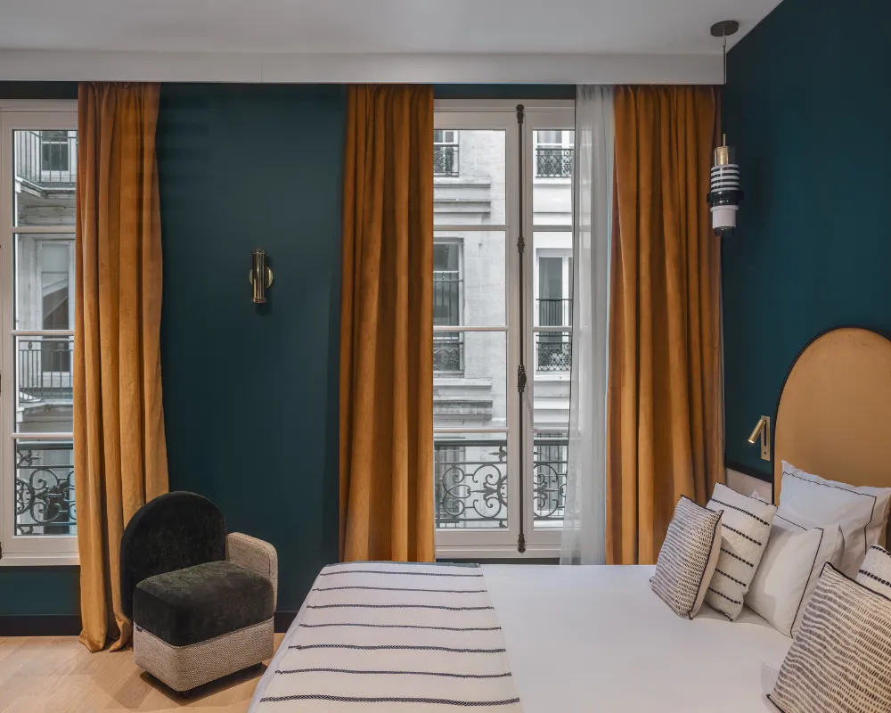 Chambre bleu paon avec fauteuil sombre, rideaux moutarde et lit à tête de lit dorée devant deux fenêtres à balcons parisiens, icône de menu en haut à droite