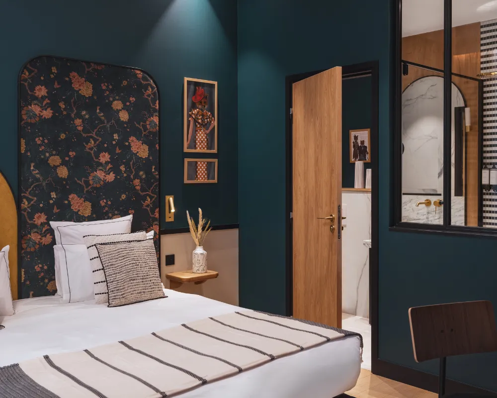 Lit à tête de lit florale sous un mur bleu paon, porte en bois ouverte sur une salle de bain à verrière