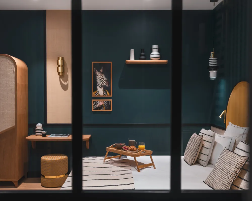 Vue à travers une verrière noire sur une chambre bleu paon avec plateau petit-déjeuner sur le lit et penderie dorée