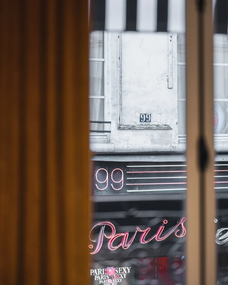 Vue entre deux rideaux moutarde sur une devanture parisienne, enseigne néon rose Paris et numéro 99 éclairés
