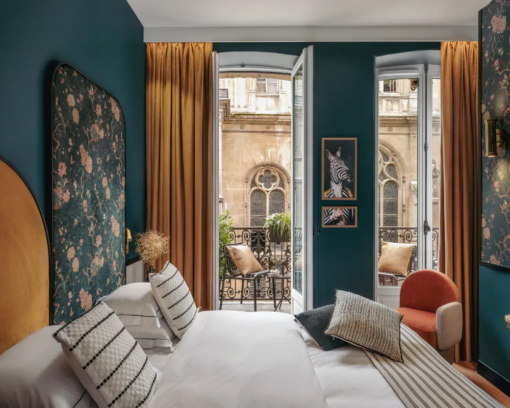 Chambre bleu paon avec tête de lit florale et rideau moutarde, double fenêtre ouverte sur un balcon face à une façade gothique