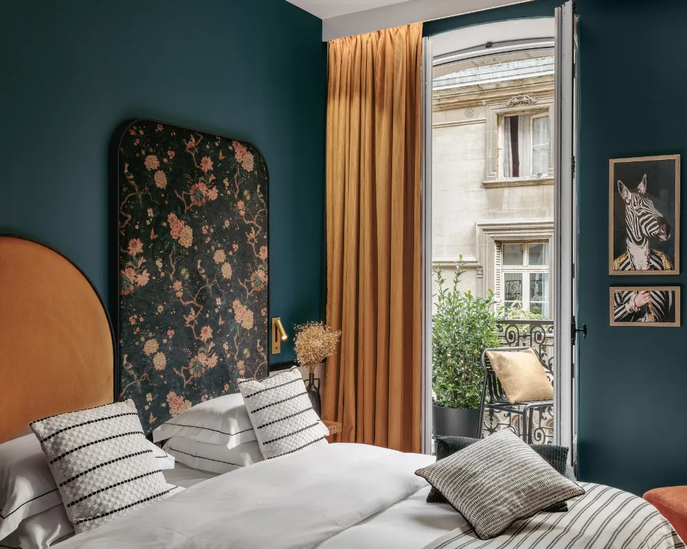 Chambre bleu paon avec tête de lit florale et arche dorée, rideau moutarde ouvert sur un balcon face à une façade parisienne