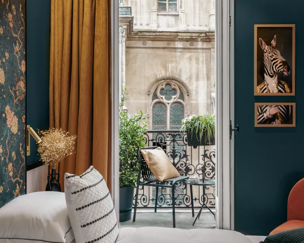 Coin lit avec coussin doré, porte-fenêtre ouverte sur un balcon en fer forgé garni de plantes face à une rosace gothique