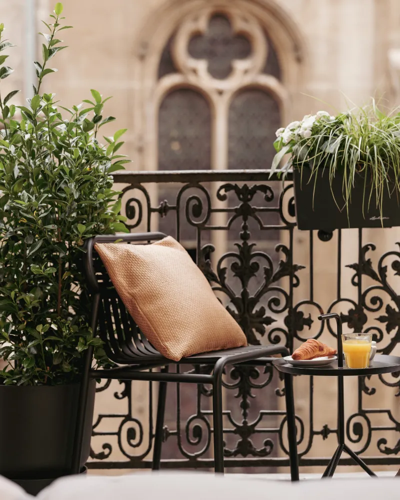 Balcon en fer forgé garni de plantes vertes avec fauteuil à coussin doré, vue sur une fenêtre à quatre-feuilles gothique