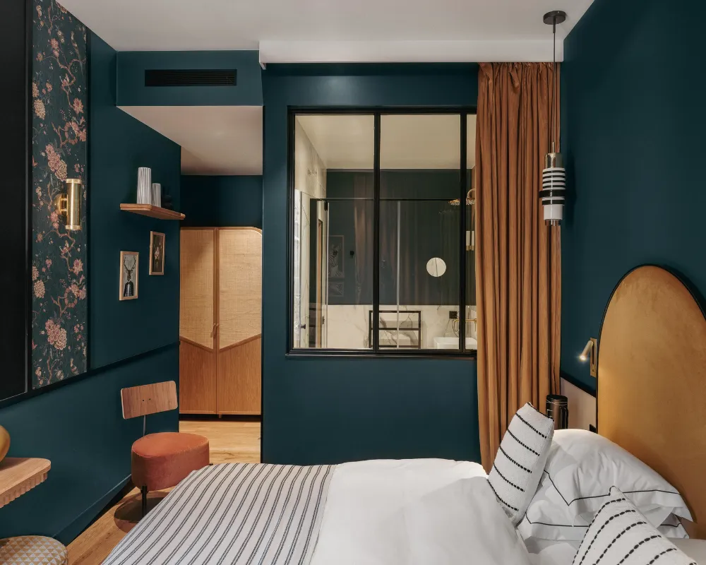 Chambre bleu paon avec tête de lit dorée arquée, verrière intérieure vitrée donnant sur une salle de bain