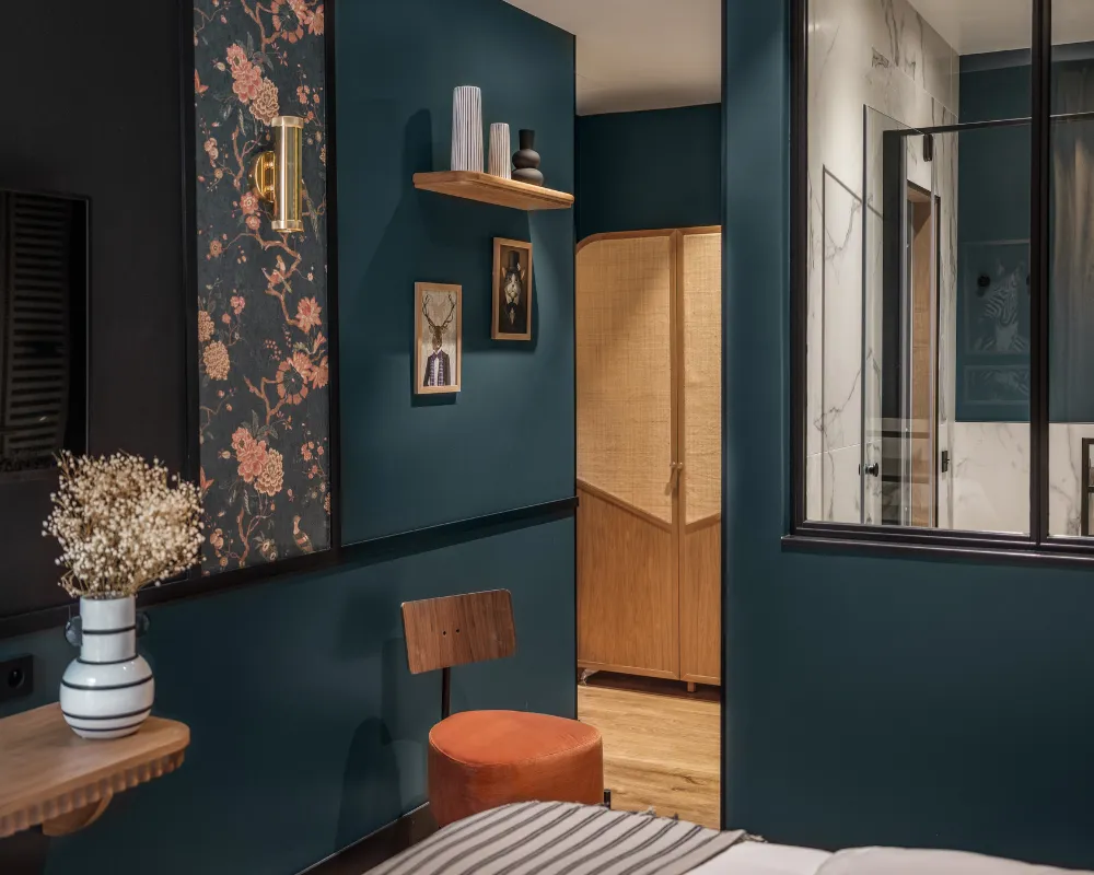 Chambre bleu paon au papier peint floral orangé, guéridon à bouquet séché et chaise camel devant une porte en bois clair