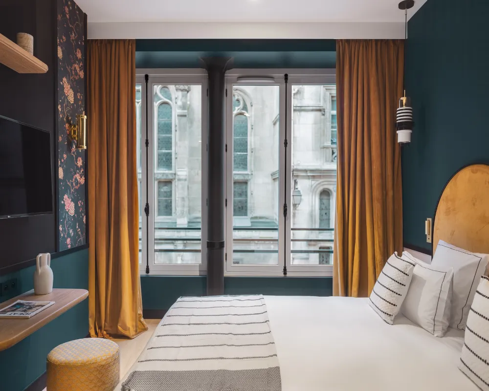 Chambre bleu paon avec lit à tête de lit dorée, rideaux moutarde et double fenêtre offrant une vue sur une façade gothique