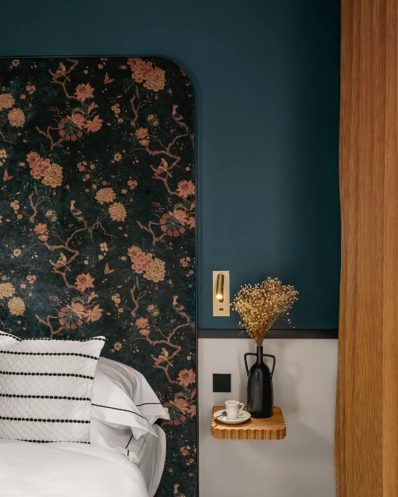 Panneau de tête de lit floral sombre sur mur bleu paon, tablette en bois avec bouquet séché et interrupteur doré, porte en bois à droite