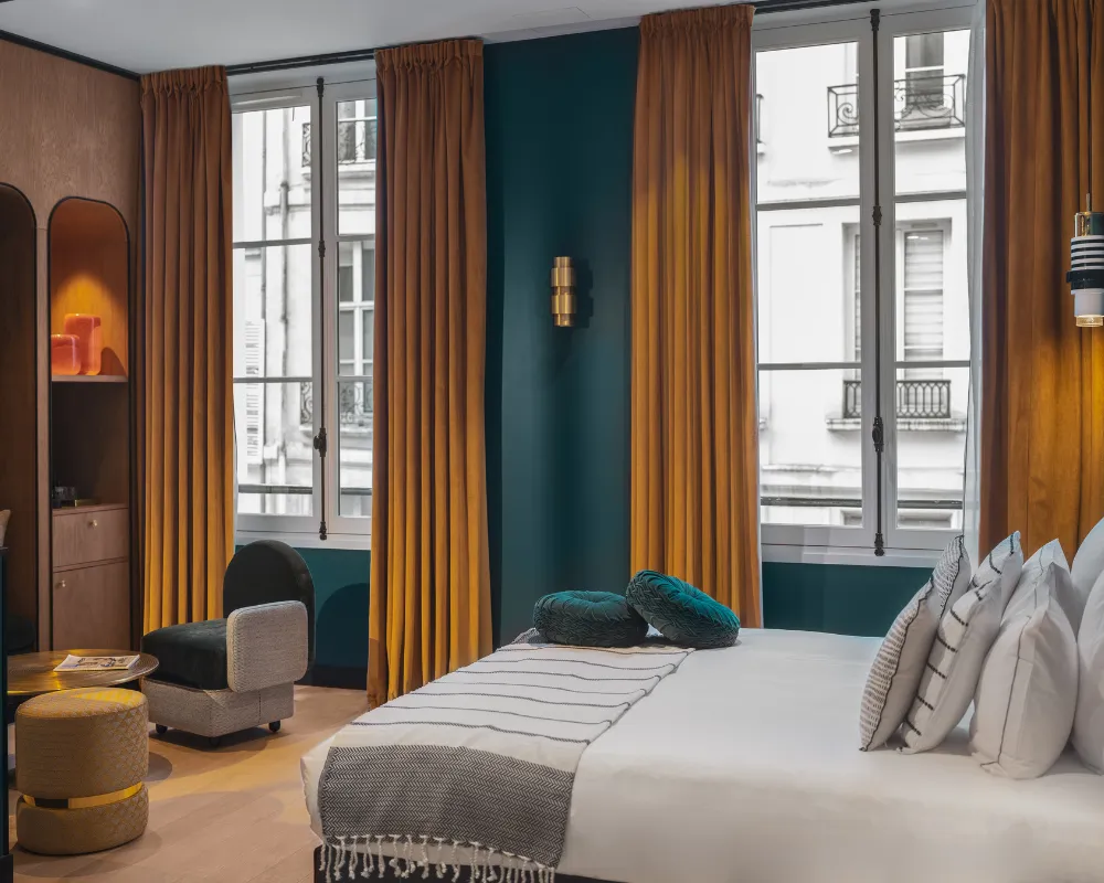Chambre bleu paon avec lit à baldaquin, rideaux dorés et fauteuils turquoise devant une fenêtre parisienne