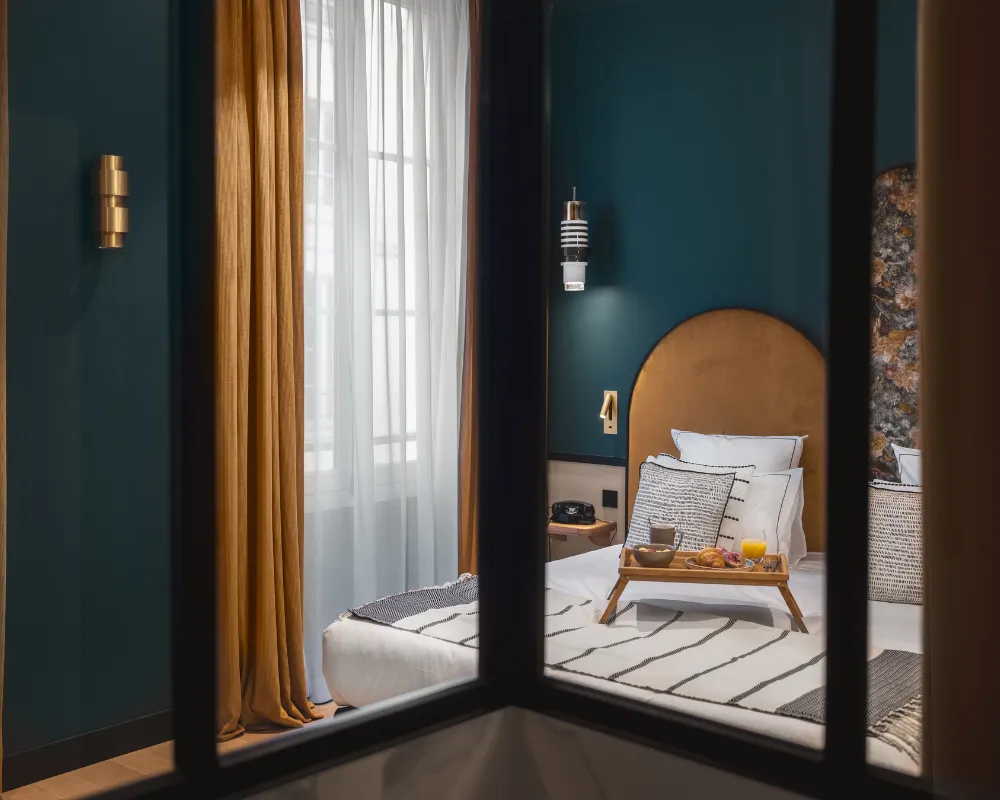 Vue à travers une fenêtre intérieure sur un lit blanc à tête de lit dorée, plateau petit-déjeuner posé dessus