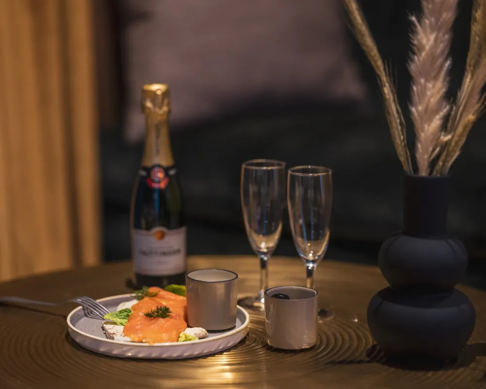Assiette de saumon garnie posée sur une table en bois, bouteille de champagne et flûtes à côté d'un vase noir à épis dorés